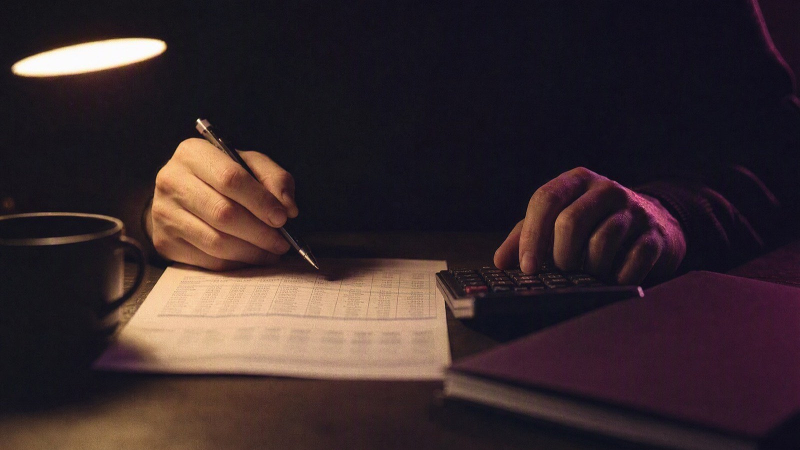 Zwei Hände am Schreibtisch: die eine mit Stift über einer Aufstellung von Zahlen, die andere am Taschenrechner